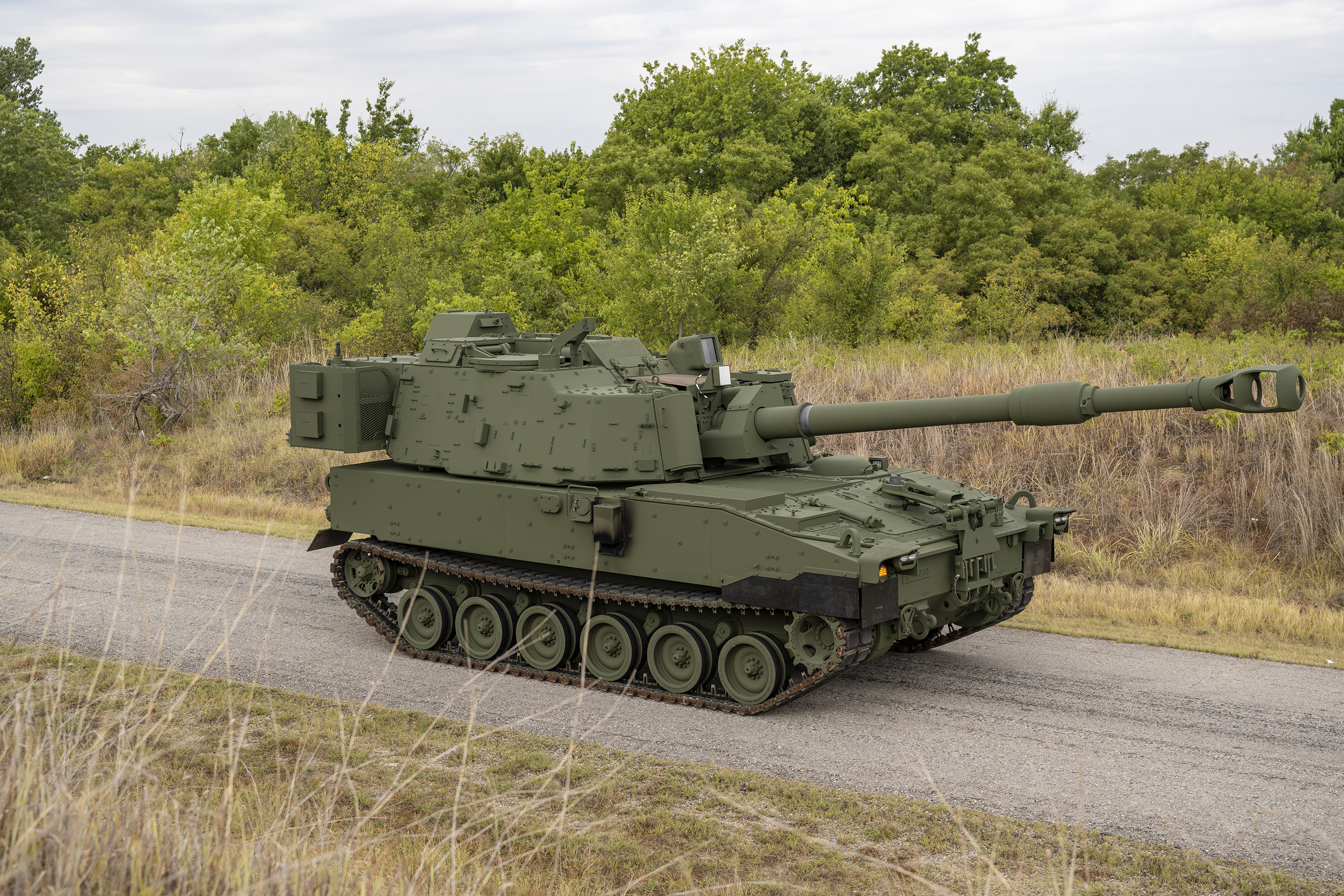A green M109A7 vehicle drives down a gravel road