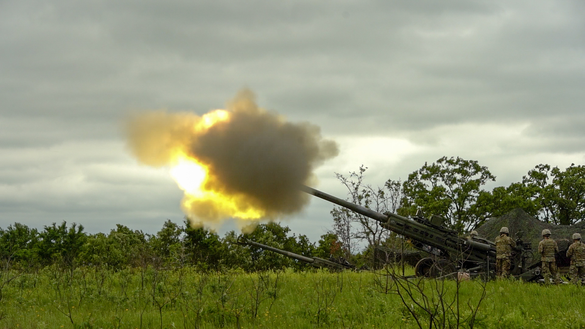 M777 firing in a grassy field on a cloudy day.