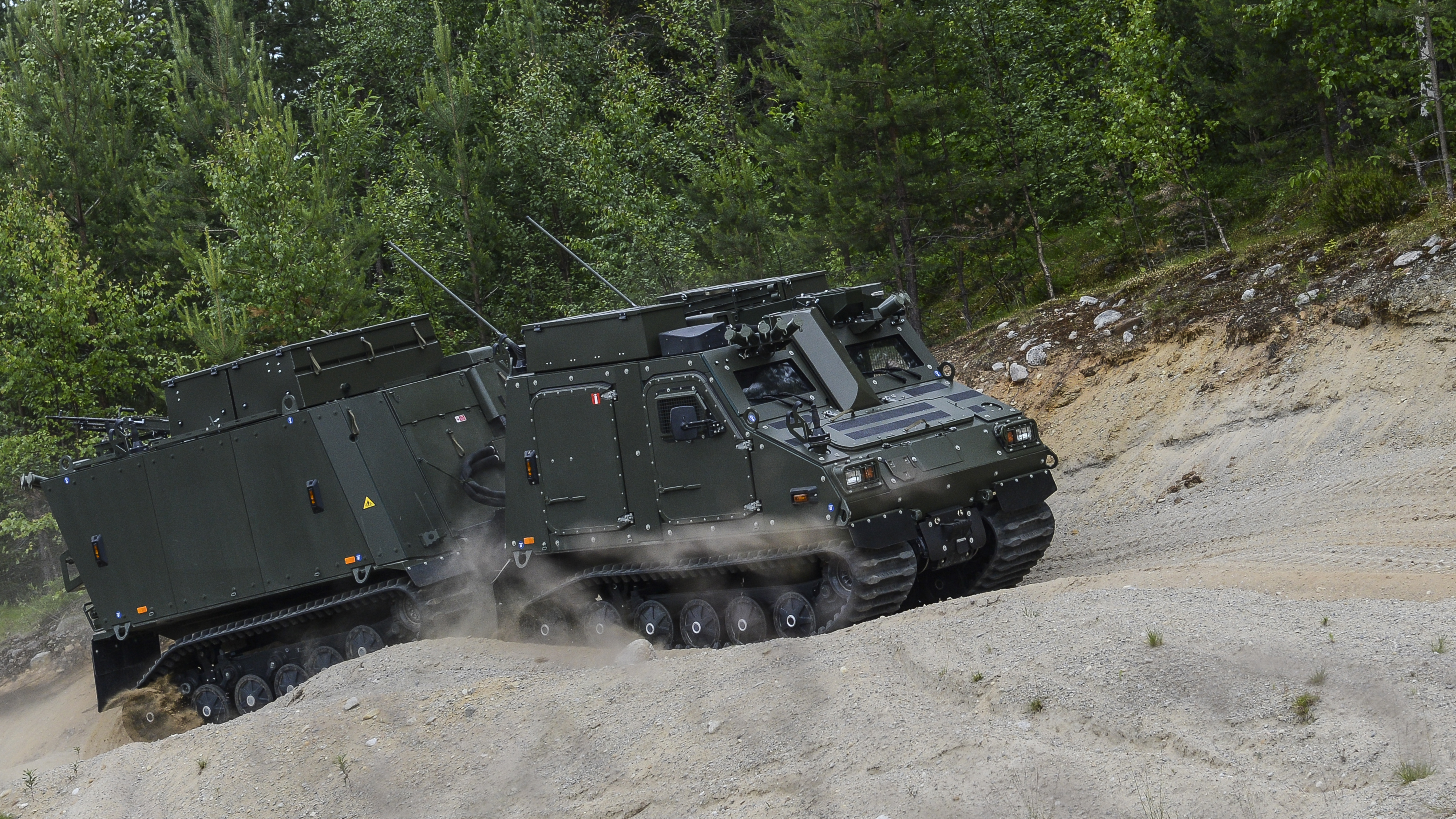A dark BvS10 driving up a gravel hill with forest in the background