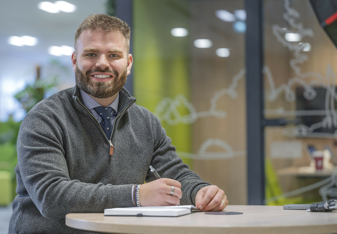 David holding a pen and paper sat in an office and looking towards the camera smiling