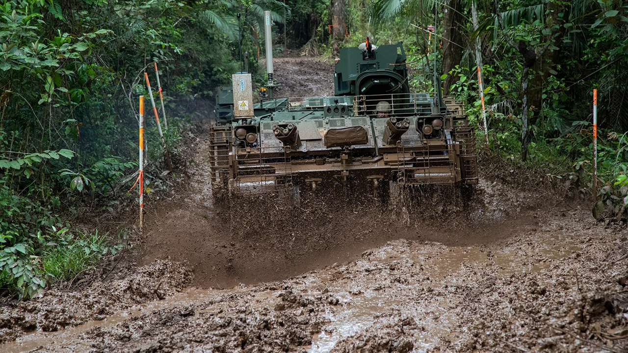 A green AMPV sprays mud as it drives through mud