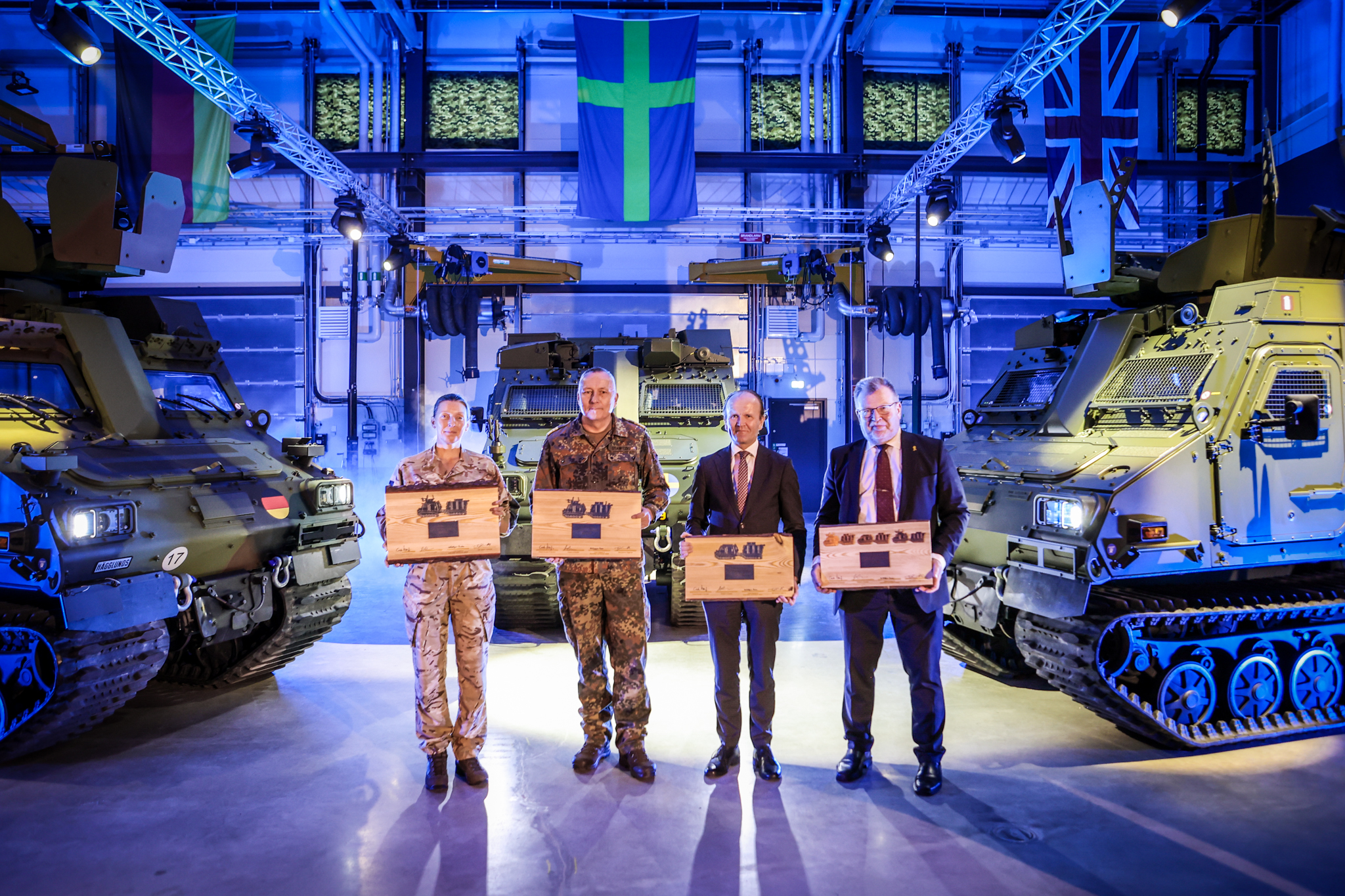 Two soldiers and two businessmen stand in from of BvS10 vehicles