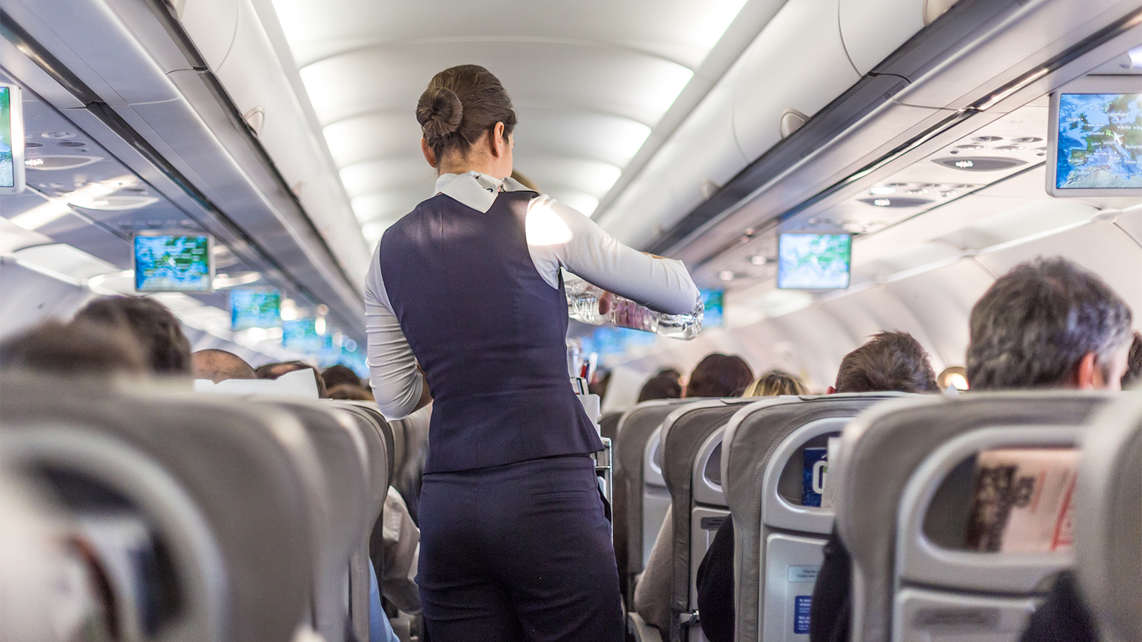 A flight attendant pours water for a passenger in an orderly, well-lit airline cabin under control via BAE Systems’ modular Cabin Management System. 